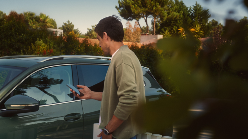 Un ragazzo si trova accanto a VW Tiguan verde scuro e aziona il sistema opzionale Park Assist Plus tramite il suo smartphone.